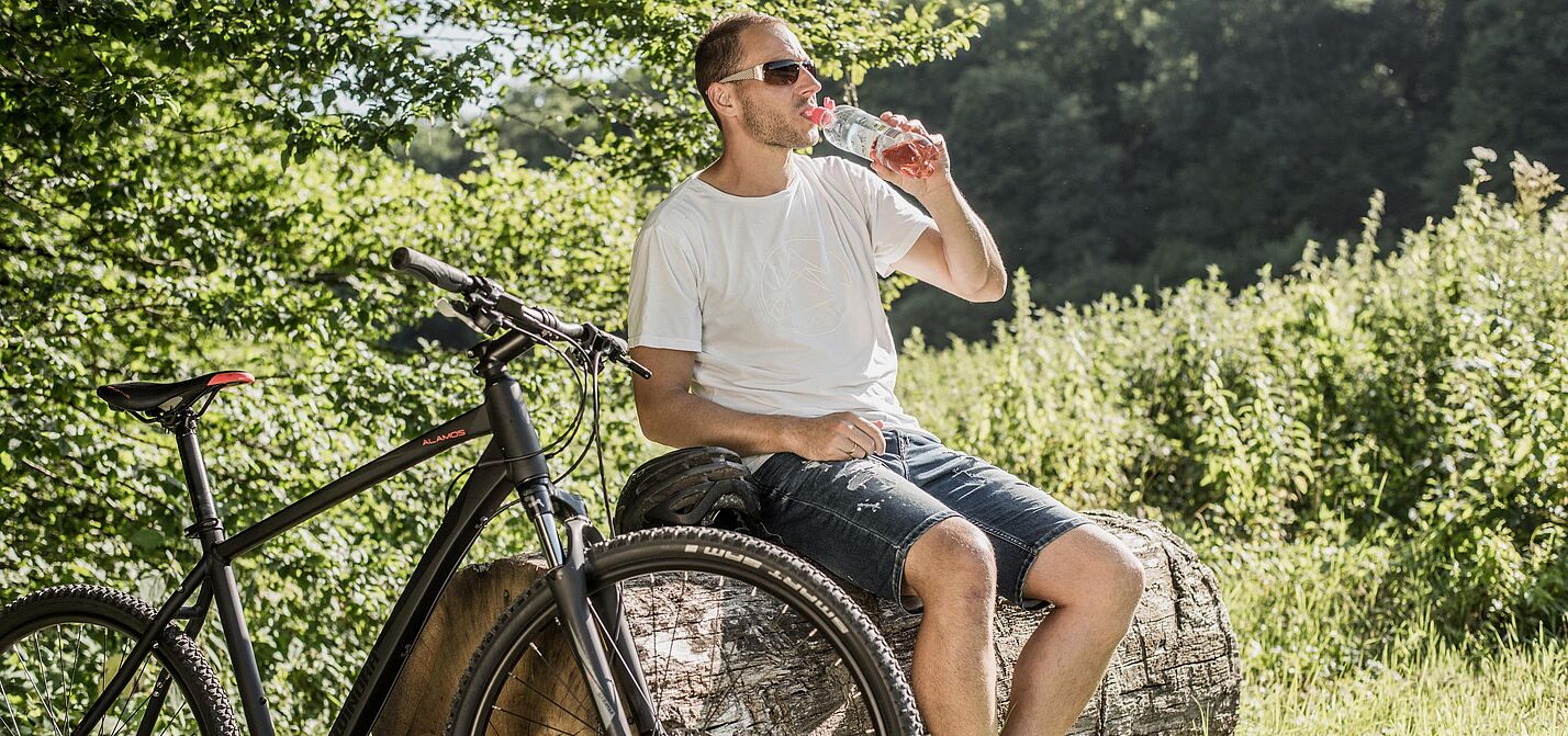 Radfahrer mit Sonnenbrille trinkt Wasser aus einer Flasche während einer Pause auf einem Baumstamm. Sein schwarzes Mountainbike steht daneben.